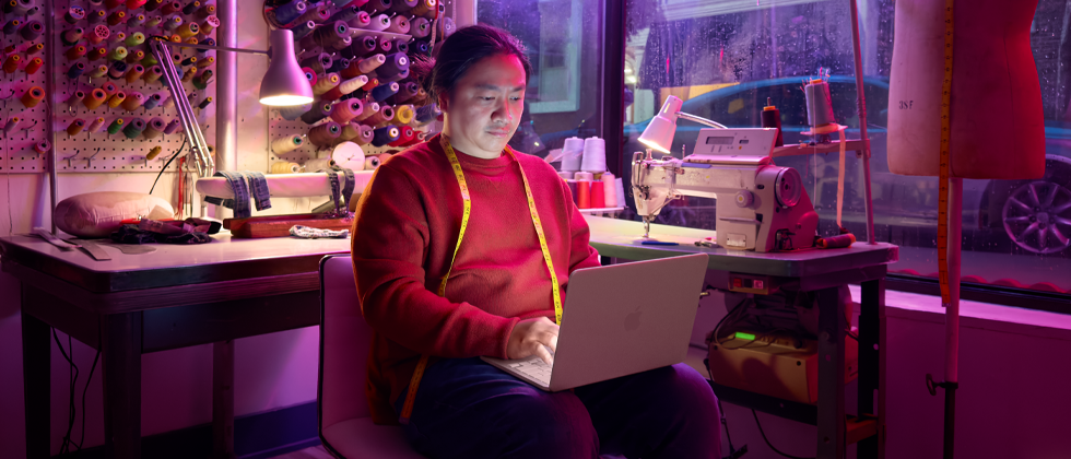 Eine Person nutzt ein MacBook Neo in Rosa auf dem Schoß, das nicht am Strom angeschlossen ist, in einem Arbeitsbereich mit einer Nähmaschine und Zubehör wie Garn und einer Schneiderpuppe