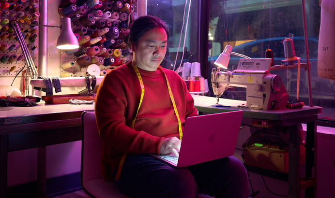 Eine Person nutzt ein MacBook Neo in Rosa auf dem Schoß, das nicht am Strom angeschlossen ist, in einem Arbeitsbereich mit einer Nähmaschine und Zubehör wie Garn und einer Schneiderpuppe